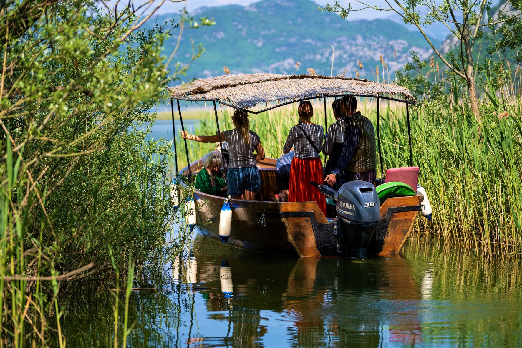 A photo shows a group of people, inside a traditional wooden boat, sailing through a channel fringed by reeds.