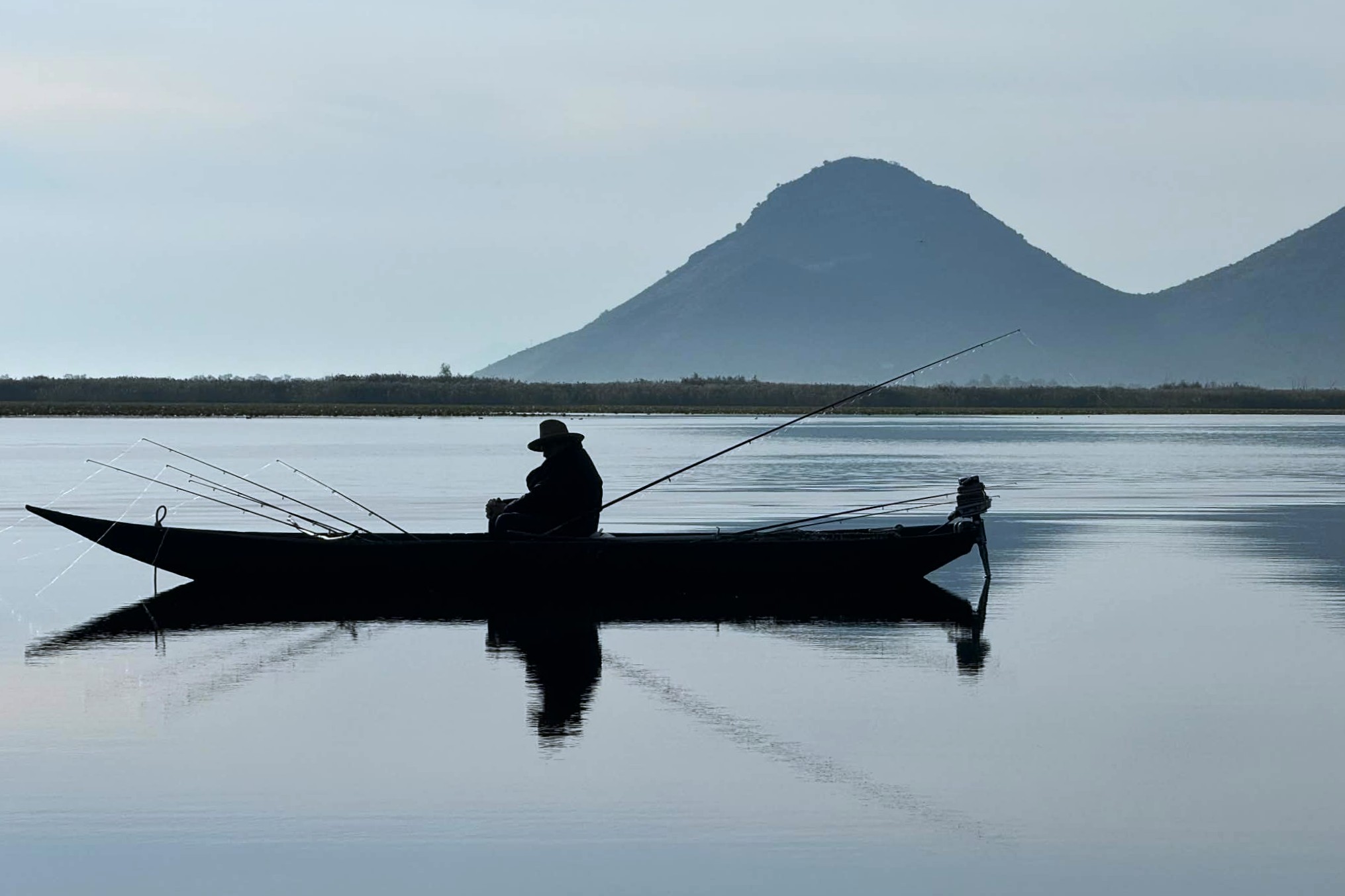 Silhouette of a fisherman sitting in a narrow wooden boat on a calm lake, surrounded by several fishing rods, with a large cone-shaped mountain in the background.
