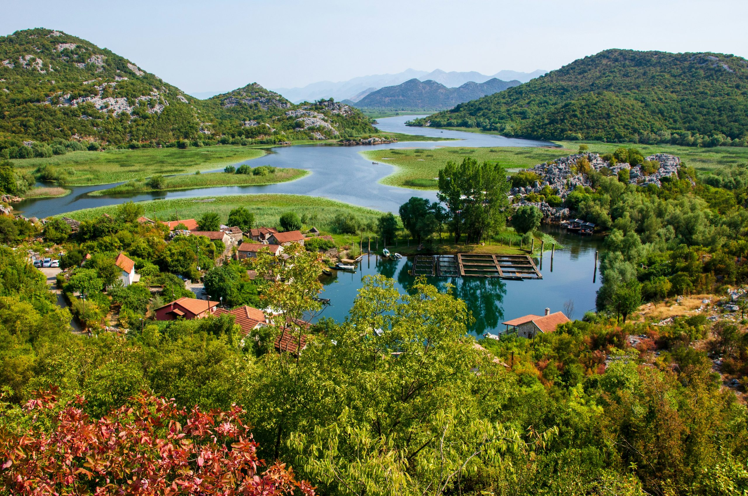A high-angle, wide landscape shot of Karuč village nestled on the shores of Lake Skadar in Montenegro. In the foreground, vibrant green foliage and red-leafed shrubs frame a small cluster of stone houses with traditional terracotta tiled roofs. A calm, blue-water bay features wooden floating structures, likely for fishing or aquaculture. Beyond the village, a winding river snakes through lush green marshlands and rolling, densely forested hills that recede into hazy blue mountains in the distance under a clear, bright sky.
