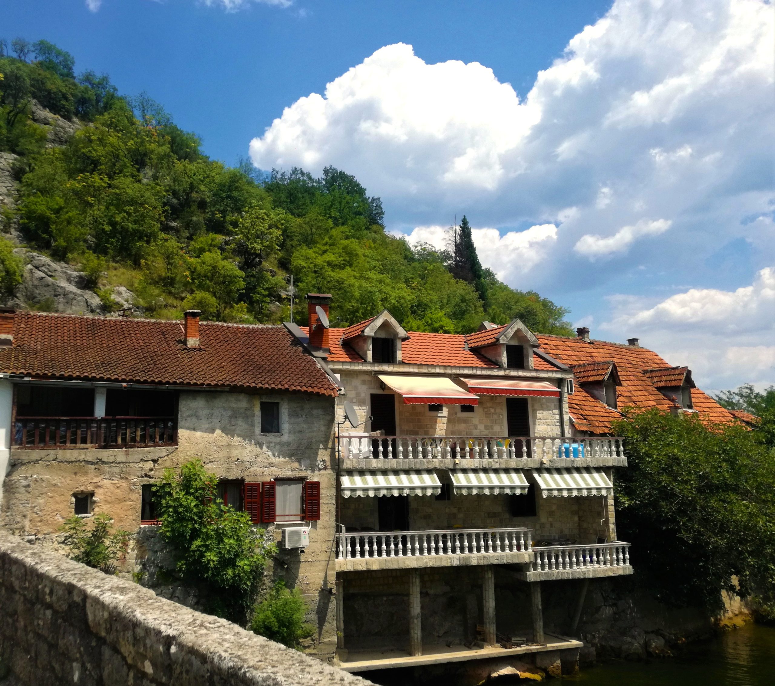 A row of historic, multi-story stone buildings with terracotta tiled roofs and wooden shutters, built directly along a lush, green hillside in Rijeka Crnojevića, Montenegro. The central house features two levels of white stone balconies with striped awnings overlooking a river.