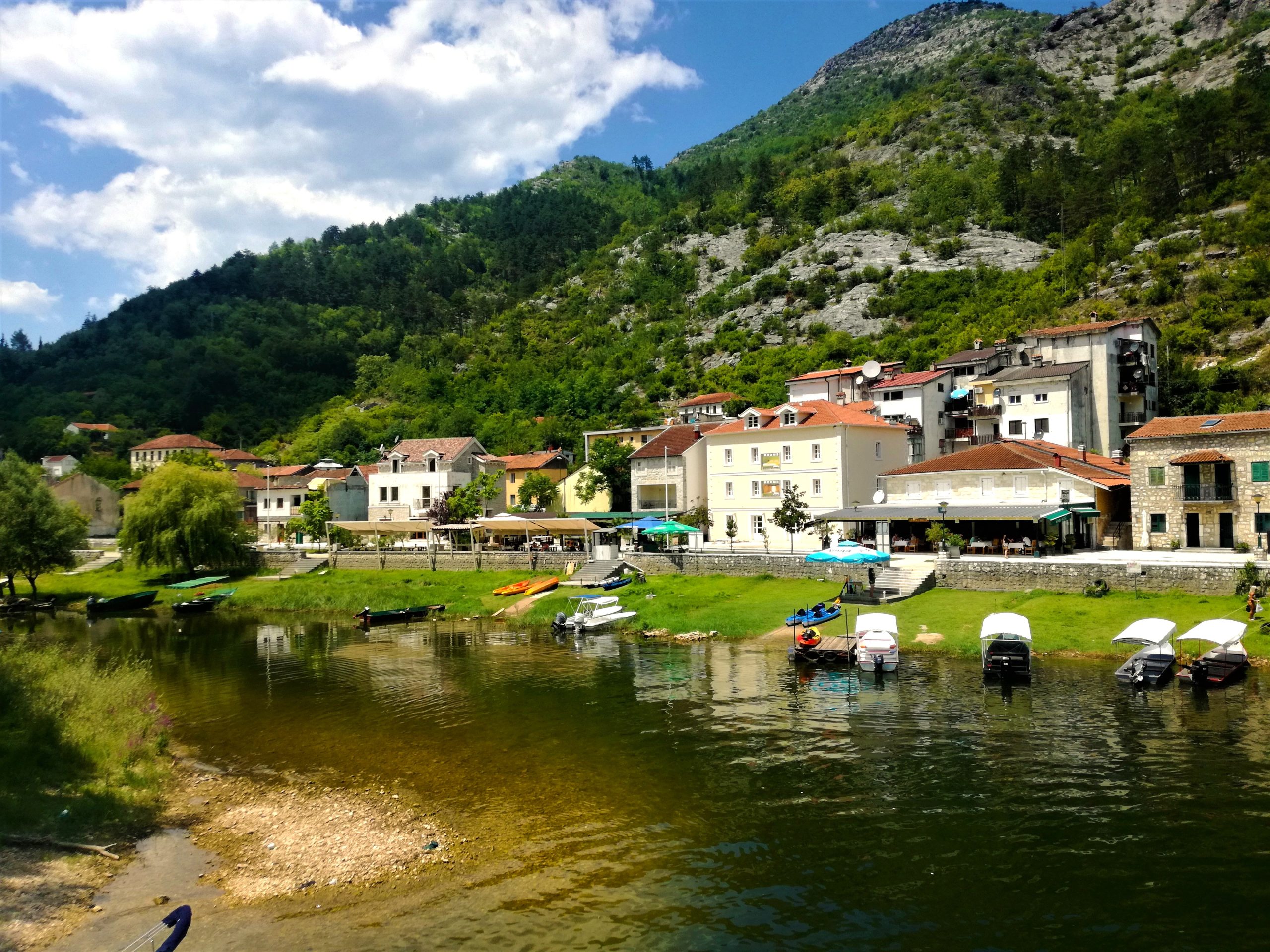 A wide, sunny view of the riverside village of Rijeka Crnojevića, Montenegro. A row of traditional stone and pastel-colored buildings with red-tiled roofs sits along a grassy bank. Several small tourist boats with white canopies are docked along the edge of the calm, dark river, which reflects the town and the lush, green mountains rising steeply in the background under a blue sky with scattered clouds.