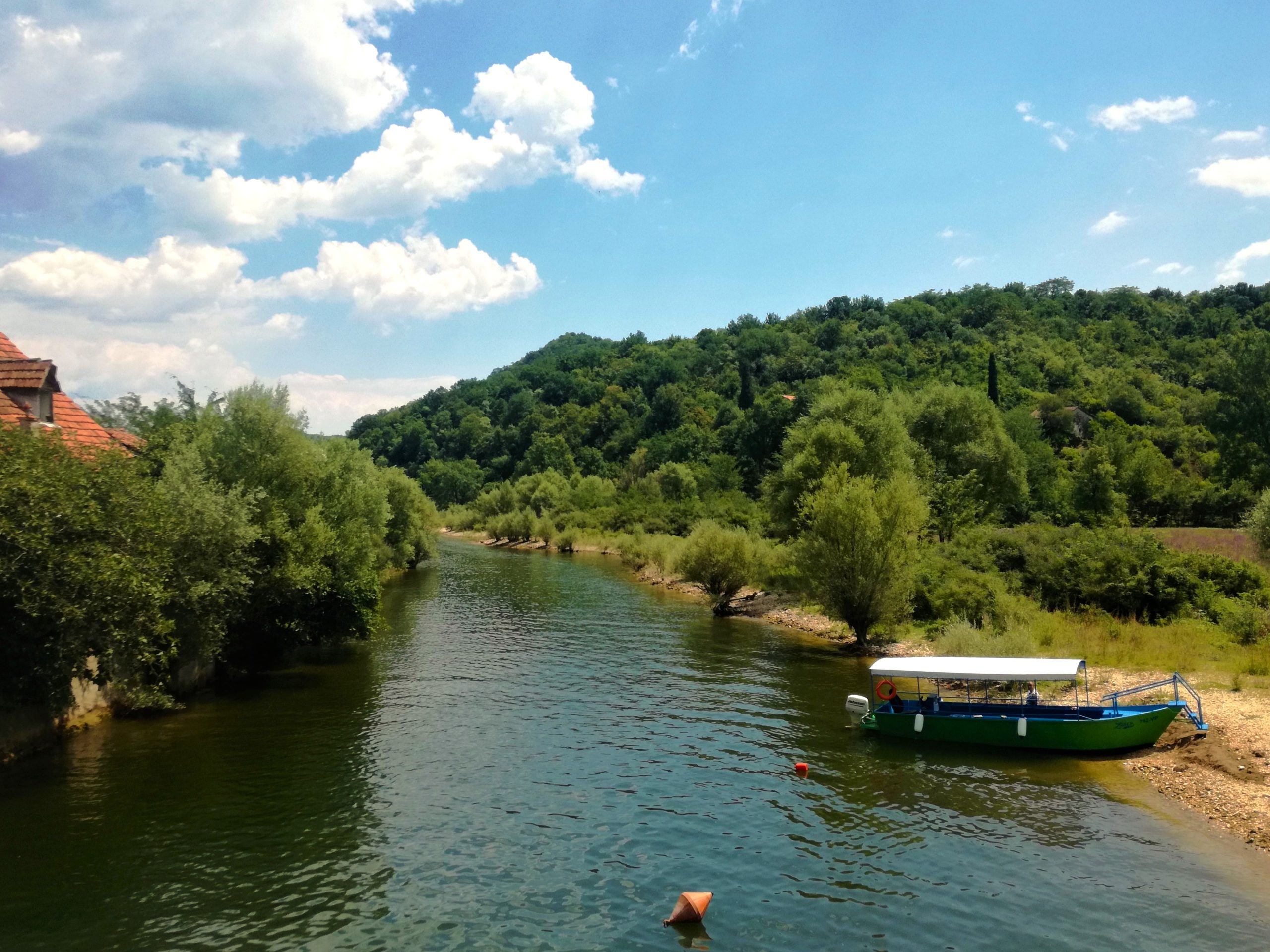 A wide, informative view of the Rijeka Crnojevića river in Montenegro. The photo shows a green wooden boat moored in the foreground on a grassy bank, with a calm river extending into the distance. The river is flanked by lush, verdant hills and dense green trees under a bright blue sky with scattered white clouds.